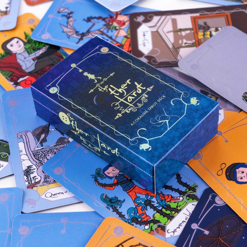 Puede incluir: Una caja rectangular para el mazo The Other Tarot, con fondo azul oscuro, texto en color dorado e ilustraciones de l&iacute;neas que representan llaves y botones. La caja est&aacute; rodeada por varias cartas de tarot con personajes y escenas ilustradas a color.