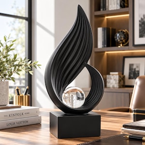 Può includere: Una scultura astratta nera caratterizzata da una struttura a coste a forma di fiamma che si curva verso l'alto e verso l'interno. Una sfera di vetro trasparente è incastonata nella curva inferiore della forma nera, montata su una base solida.