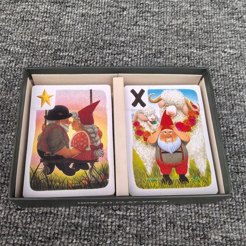 K&ouml;nnte beinhalten: Eine dunkelgraue Schachtel mit zwei Spielkarten. Die linke Karte zeigt einen gelben Stern, ein Gnom-Paar auf einer Schaukel und einen Sonnenuntergang. Die rechte Karte zeigt den Buchstaben X, einen Gnom mit wei&szlig;em Bart und zwei wei&szlig;e Schafe.