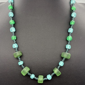 May include: A necklace featuring alternating frosted green and light blue beads interspersed with small, dark metallic spacers. The beads vary in shape, including rounded and rectangular forms, strung together to create a continuous circular pattern.