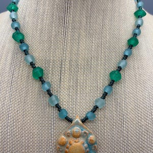 May include: A necklace featuring a central teardrop-shaped ceramic pendant in tan and blue with a raised circular pattern. The pendant is suspended from a strand of alternating translucent green and light blue glass beads separated by small black spacers.