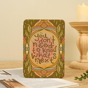 Peut inclure: Une impression rectangulaire avec une bordure vert olive ornée de motifs de feuilles et d'yeux stylisés. Le centre présente un dégradé orange et rose avec le texte brun foncé : You don't need to know what's next.