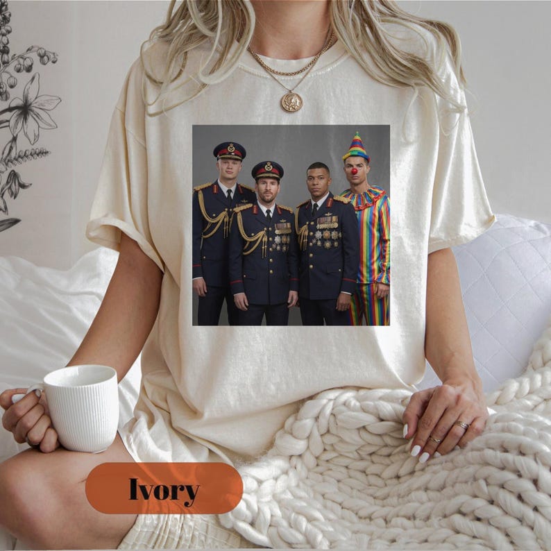 Op de afbeelding: Een ivoorkleurig t-shirt met een grafische print van drie personen in donkere militaire uniformen met gouden accenten en medailles, naast een vierde persoon in een kleurrijk gestreept clownskostuum met een feesthoedje.