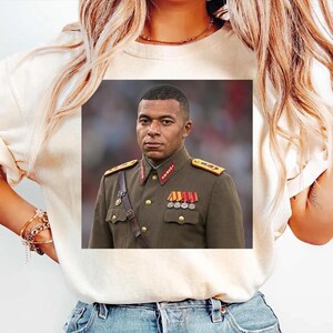 K&ouml;nnte beinhalten: Ein cremefarbenes T-Shirt mit einem mittigen Grafikdruck einer Person in einer dunkelolivgr&uuml;nen Uniform im Milit&auml;rstil mit goldenen Kn&ouml;pfen, Schulterklappen sowie einer Brustdekoration aus bunten Dienstb&auml;ndern und Orden.