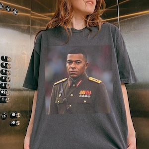 K&ouml;nnte beinhalten: Ein anthrazitfarbenes Kurzarm-T-Shirt mit einem Grafikdruck einer Person in einer formellen Uniform im Milit&auml;rstil mit goldenen Schulterklappen, roten Kragenspiegeln und mehreren Medaillen an der olivgr&uuml;nen Jacke.