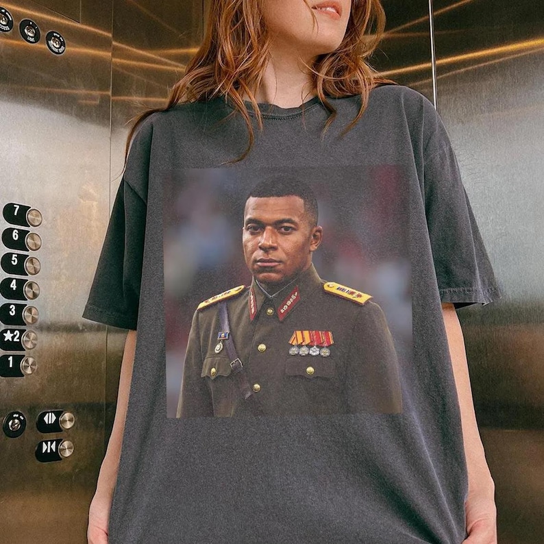 K&ouml;nnte beinhalten: Ein anthrazitfarbenes Kurzarm-T-Shirt mit einem Grafikdruck einer Person in einer formellen Uniform im Milit&auml;rstil mit goldenen Schulterklappen, roten Kragenspiegeln und mehreren Medaillen an der olivgr&uuml;nen Jacke.