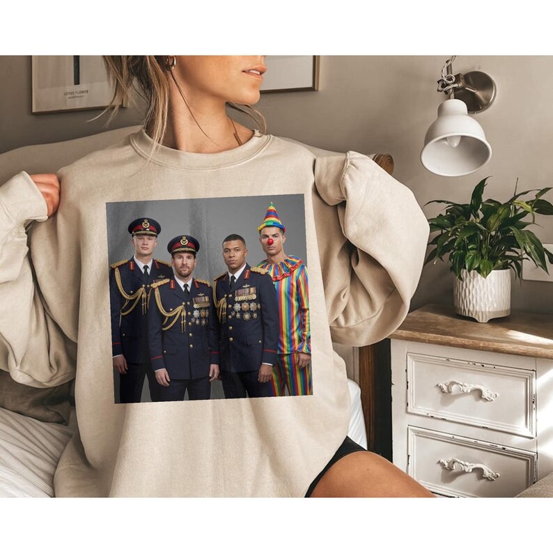 Op de afbeelding: Een beige sweater met ronde hals en een grafische print van vier personen. Drie figuren dragen donkerblauwe militaire uniformen met gouden accenten en medailles, terwijl de vierde figuur een kleurrijk gestreept clownskostuum draagt.