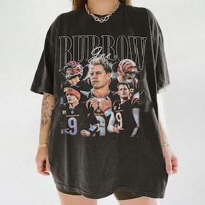 May include: A charcoal gray short-sleeve t-shirt featuring a graphic print of football player Joe Burrow in various poses. The text BURROW is printed in large white letters at the top, with the name Joe written in a script font underneath the text.