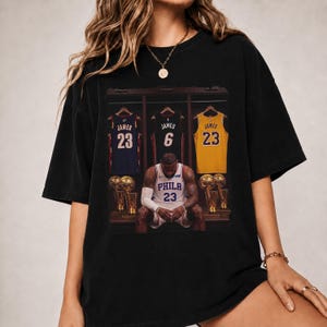 May include: A black short-sleeve t-shirt featuring a graphic print of a basketball player wearing a white jersey with the text PHILA 23. Behind the player are three hanging jerseys with the name JAMES and numbers 23, 6, and 23, plus gold trophies.