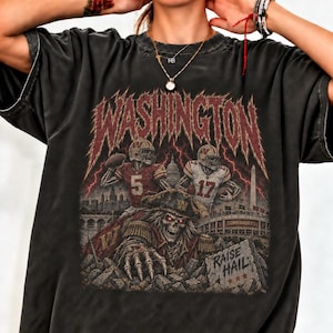 May include: A charcoal gray short-sleeve t-shirt featuring a graphic print with the text WASHINGTON and RAISE HAIL. The design includes two football players in jerseys numbered 5 and 17, a skeleton in a military hat, and a city skyline background.