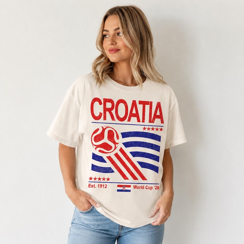 Pode incluir: Uma t-shirt de manga curta em tom off-white com uma estampa gr&aacute;fica em vermelho e azul. O texto diz CROATIA, Est. 1912 e World Cup '26. O design inclui uma bola de futebol estilizada, cinco estrelas vermelhas e uma pequena bandeira croata.