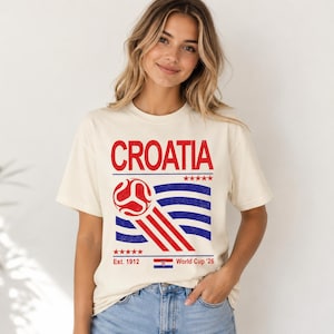 Pode incluir: Uma t-shirt de manga curta cor de creme com um gr&aacute;fico que apresenta o texto vermelho CROATIA sobre uma bola de futebol vermelha estilizada e linhas onduladas azuis. O design inclui cinco estrelas vermelhas, o texto Est. 1912, uma bandeira croata e World Cup '26.