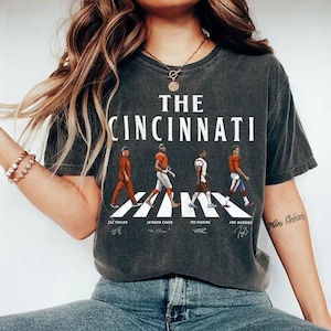 May include: A charcoal gray short-sleeve t-shirt featuring the text THE CINCINNATI above a graphic of four football figures walking across a crosswalk. Below each figure are the names Zac Taylor, Ja'Marr Chase, Tee Higgins, and Joe Burrow.