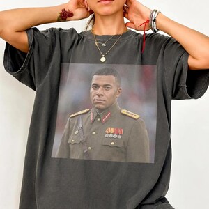 K&ouml;nnte beinhalten: Ein anthrazitfarbenes Kurzarm-T-Shirt mit einem gro&szlig;en rechteckigen Grafikdruck auf der Brust. Der Druck zeigt eine Person in einer dunkelolivgr&uuml;nen Milit&auml;runiform mit goldenen Schulterklappen und mehreren bunten Medaillen.