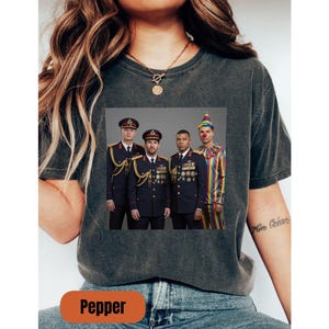 Op de afbeelding: Een antracietgrijs T-shirt met korte mouwen en een grafische print van drie personen in donkerblauwe militaire uniformen met gouden accenten en medailles, naast een persoon in een kleurrijk gestreept clownskostuum met feesthoed.