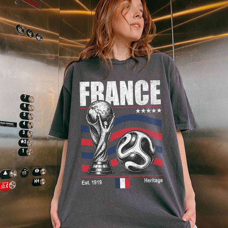 Puede incluir: Una camiseta de manga corta gris carb&oacute;n con un estampado gr&aacute;fico que incluye la palabra FRANCE en letras blancas. El dise&ntilde;o presenta un trofeo de f&uacute;tbol, un bal&oacute;n, cinco estrellas, l&iacute;neas onduladas rojas y azules, una peque&ntilde;a bandera francesa y el texto Est. 1919 Heritage.