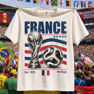 Puede incluir: Una camiseta de manga corta de color crema con un estampado gr&aacute;fico que incluye la palabra FRANCE en azul, un trofeo de f&uacute;tbol, un bal&oacute;n de f&uacute;tbol, cinco estrellas azules y el texto Est. 1919 Heritage con una peque&ntilde;a bandera francesa.