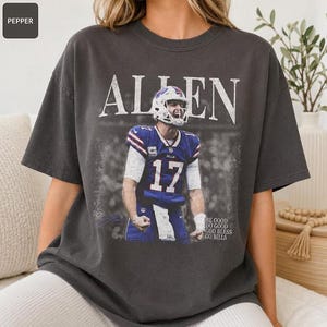 May include: A charcoal gray short-sleeve t-shirt featuring a graphic of a football player in a blue jersey with the number 17. The text "ALLEN" appears in large white letters above the player, and "BE GOOD DO GOOD GOD BLESS GO BILLS" is printed below.