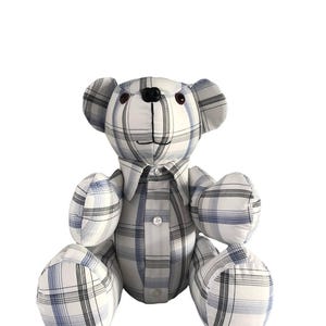 May include: A plaid teddy bear made from a white, blue, and black checked shirt. The bear has a black nose, brown eyes, and a button-down shirt collar. The bear is sitting with its arms and legs outstretched.