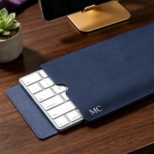 Può includere: Una custodia per tastiera in pelle blu navy con finitura liscia e le iniziali MC impresse nell'angolo inferiore. La custodia è parzialmente aperta e rivela una tastiera wireless bianca con tasti argentati all'interno.