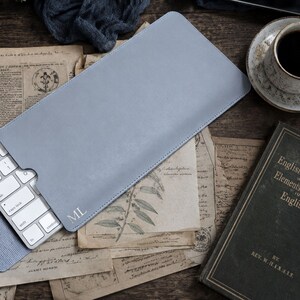 Peut inclure: Une housse de clavier rectangulaire bleu clair avec une finition lisse et les initiales ML gaufrées dans le coin inférieur. La housse est partiellement ouverte, révélant un clavier sans fil blanc aux touches métalliques à l'intérieur.