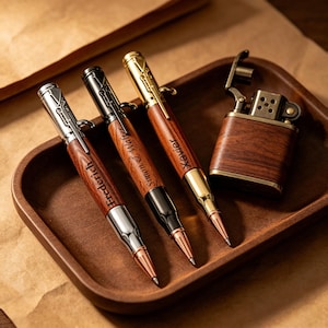 May include: A wooden tray holds three engraved pens and a lighter. The pens have a wood grain finish with silver, black, and gold accents. Each pen is engraved with a name. The lighter has a wood grain finish and a metal top.