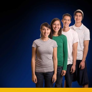 May include: Four people stand in a row, smiling at the camera. The first person wears a gray t-shirt and jeans. The second wears a green long-sleeve shirt and jeans. The third wears a white t-shirt and jeans. The fourth wears a striped shirt, beanie, and shorts.