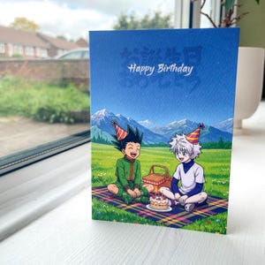May include: A rectangular greeting card featuring a blue sky with the text Happy Birthday. Below, two anime characters with party hats sit on a plaid blanket in a green field with mountains in the background, next to a picnic basket and a cake.