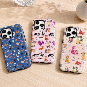 May include: Three smartphone cases featuring repeating patterns of various cartoon cats. The left case is blue, the center case is light pink, and the right case is cream-colored. Each case displays multiple small, colorful feline illustrations.