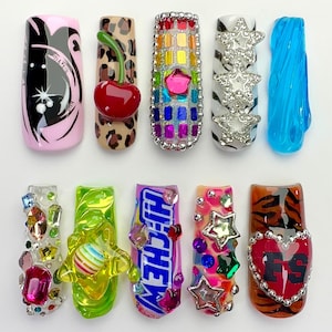 Puede incluir: Diez uñas postizas con diseños variados: un gráfico rosa y negro, estampado de leopardo con una cereza 3D, una cuadrícula arcoíris con una estrella, rayas de cebra blancas con estrellas plateadas, ondas azules translúcidas, gemas, un remolino verde con una esfera a rayas, un envoltorio de caramelo Hi-Chew, estrellas coloridas y un estampado de tigre con un corazón.