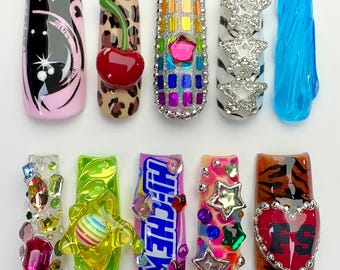 May include: Ten press-on nails featuring diverse designs: a pink and black graphic, a leopard print with a 3D cherry, a rainbow grid with a star, white zebra stripes with silver stars, blue translucent waves, clear gems, a green swirl with a striped sphere, a Hi-Chew candy wrapper, colorful stars, and a tiger print with a heart.