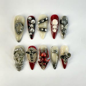 May include: A collection of ten long, almond-shaped artificial nails. Each nail features unique embellishments, including silver-toned charms, beads, and abstract designs. The color palette includes cream, red, and silver accents, creating a bold and artistic aesthetic.