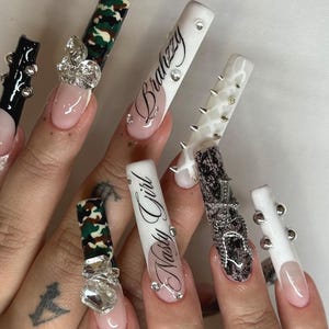 Puede incluir: Primer plano de uñas artificiales largas y adornadas. Los diseños incluyen camuflaje, pedrería, encaje y las palabras "Brahzzy" y "Nasty Girl". Algunas uñas presentan tachuelas plateadas. Las uñas tienen una combinación de colores rosa y blanco.