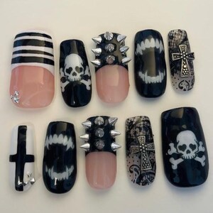 May include: A set of ten press-on nails featuring black and white designs including horizontal stripes, white skulls and crossbones, silver metal spikes, white fangs, black lace patterns with silver cross charms, and a black cross on a white base.