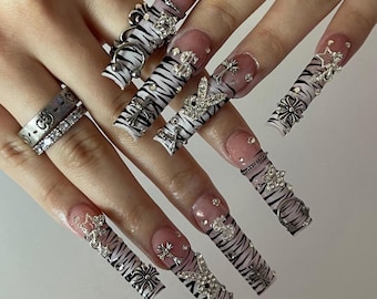 May include: Long square-shaped artificial nails featuring a white base with black tiger stripes. The nails are decorated with various silver-toned metal charms including stars, dollar signs, crosses, and rabbit silhouettes, accented with small clear rhinestones.