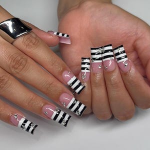 Op de afbeelding: Lange, vierkante kunstnagels met een nude roze basiskleur en horizontale zwarte en witte strepen op de uiteinden. Elke nagel is versierd met kleine, doorzichtige, reflecterende strass-steentjes langs het streeppatroon en de nagelbasis.