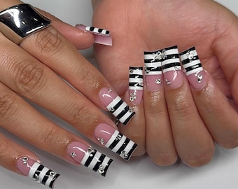 May include: Long square-shaped artificial nails featuring a nude pink base color with horizontal black and white stripes on the tips. Each nail is decorated with small, clear, reflective rhinestones placed along the striped pattern and base.