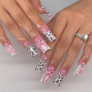 Puede incluir: Uñas postizas largas de forma cuadrada con base blanca, rayas de cebra negras y manchas de leopardo rosas. El diseño incluye lazos rosas tridimensionales, pequeñas flores rosas, dijes en forma de corazón y pedrería transparente.