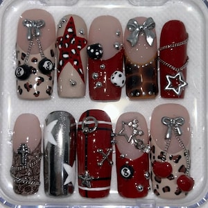 Pode incluir: Um conjunto de dez unhas postiças com vários designs, incluindo estampa de leopardo, estrelas vermelhas, dados, laços prateados, correntes, bolas de bilhar número 8, cruzes e acabamentos metálicos, dispostas num estojo de plástico transparente.