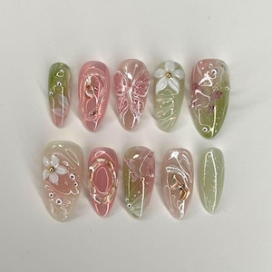 Puede incluir: Un conjunto de diez uñas artificiales con varios diseños. Las uñas presentan una mezcla de bases rosas, verdes y transparentes, adornadas con patrones florales y abstractos, detalles dorados y adornos florales en 3D. Las uñas son de forma ovalada.
