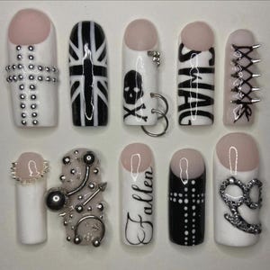 Puede incluir: Un conjunto de diez uñas postizas con varios diseños. Las uñas presentan una base blanca con detalles en negro, incluyendo una Union Jack, una calavera y la palabra "SWAG". Algunas uñas tienen tachuelas plateadas, púas y un diseño de cruz.
