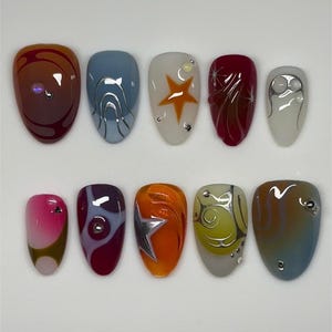 Puede incluir: Un juego de diez uñas postizas con diversos diseños que incluyen remolinos naranjas y rojos, azul con líneas plateadas, una uña blanca con una estrella naranja, rojo intenso con detalles de estrellas y blanco con adornos plateados y patrones.