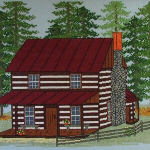 May include: A cross-stitch pattern of a brown log cabin with a red roof and a stone chimney. The cabin has a porch with a railing and a barrel on the left side. There are green trees in the background.