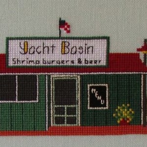 May include: A cross-stitch embroidery depicting a green building with a red roof and a sign that reads "Yacht Basin Shrimp burgers & beer". There are two people standing outside the building, and two boats docked on the right side of the image.