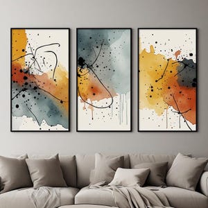 May include: A set of three vertical wall art prints featuring abstract watercolor washes in shades of mustard yellow, burnt orange, and slate blue. Each print includes black ink splatters and thin, hand-drawn black lines on a cream background.