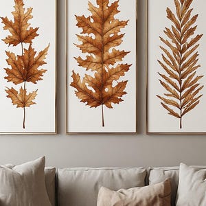Peut inclure: Un ensemble de trois cadres muraux verticaux dorés fins, chacun présentant une illustration aquarelle détaillée d'une feuille d'automne brune sur un fond blanc uni, disposés côte à côte dans un agencement uniforme.