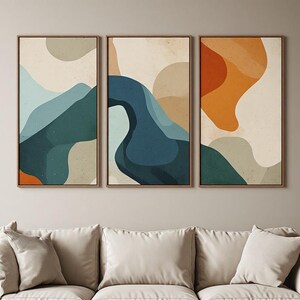 Peut inclure: Un ensemble de trois impressions d'art mural rectangulaires verticales dans des cadres en bois fins. L'œuvre présente des formes abstraites, fluides et organiques dans une palette de bleu sarcelle, bleu profond, orange, beige et crème.