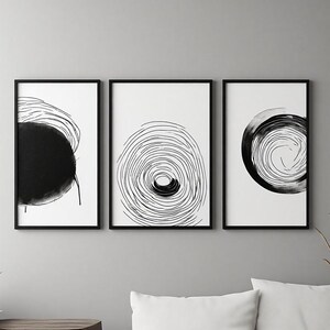 Op de afbeelding: Een set van drie ingelijste wandkunstwerken met zwart-witte abstracte ontwerpen. Elk rechthoekig frame bevat een unieke compositie van cirkelvormige inktlijnen en effen zwarte vormen op een witte achtergrond, horizontaal naast elkaar geplaatst.