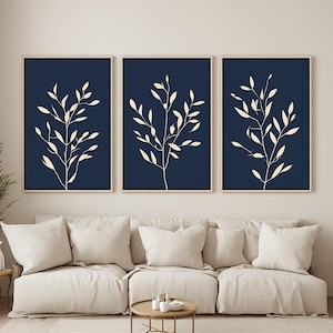 Peut inclure: Un ensemble de trois impressions murales rectangulaires présentant des illustrations de feuilles botaniques couleur crème centrées sur des fonds bleu marine foncé, chacune encadrée d'un cadre en bois clair, disposées en rangée.