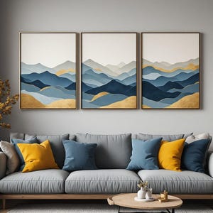 Könnte beinhalten: Ein dreiteiliges Wandbild-Set bestehend aus drei gerahmten Drucken abstrakter Gebirgszüge in Blau-, Grau- und Goldtönen. Die Kunstwerke zeigen geschichtete Gipfel mit einem sanften Verlaufseffekt, eingefasst in schmale, schlichte Rahmen.